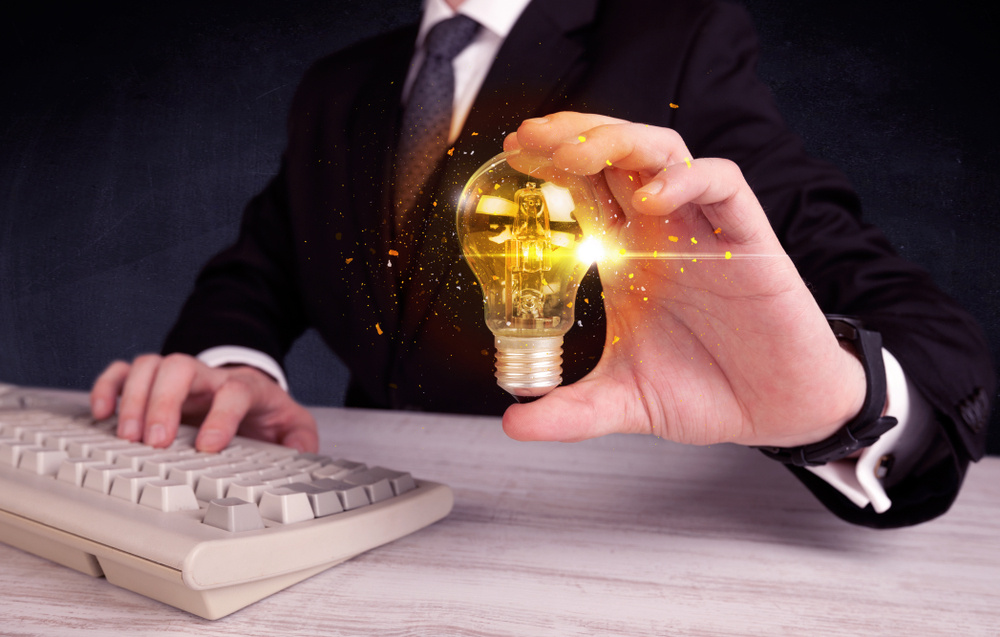 An elegant office worker holding a yellow sparkling light bulb in his hand while working in front of dark blue background concept.