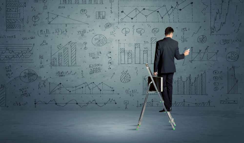 A businessman in modern stylish elegant suit standing on a small ladder and drawing pie and block charts on grey wall background with exponential progressing curves, lines, circles, blocks, numbers